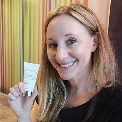 Woman smiling indoors holding a Glowbiotics Tinted Sunscreen SPF 30 tube.