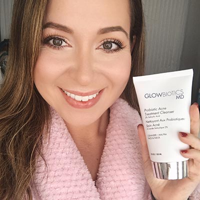Woman with pink sweater holding Glowbiotics cleanser tube, smiling at camera