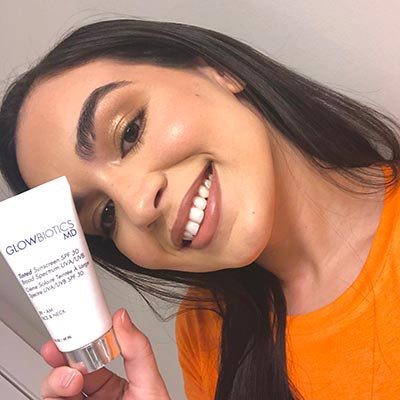 Woman in orange top smiling and holding a Glowbiotics Revitalizing Cleanser tube.